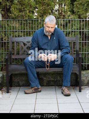 Man with grey hair and full beard, 50-59 years old, sitting on a bench on the terrace outside and praying the Rosary Stock Photo