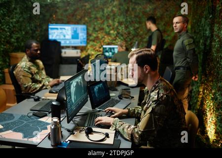 Side view of young male military officer typing on keyboard and looking at computer screen with graphic data among other specialists Stock Photo
