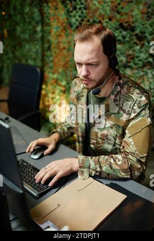 Young serious male military officer in headset keeping hand on computer keyboard and looking at screen with graphic data by workplace Stock Photo