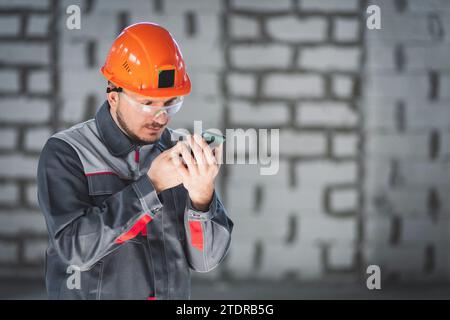 Caucasian male builder in overalls and helmet uses smartphone as radio for communication at construction site Stock Photo