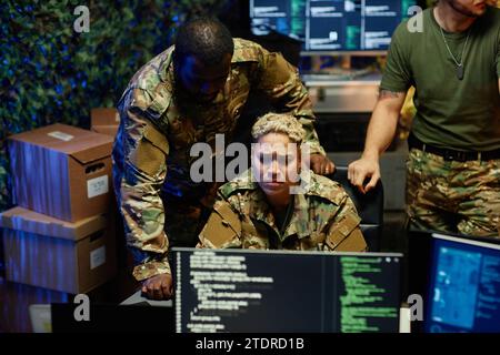 Serious female officer in camouflage uniform looking at computer screen while sitting between two young intercultural male colleagues Stock Photo