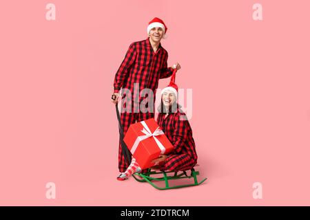Happy young couple in Christmas pajamas, with sledge and gift on pink background Stock Photo