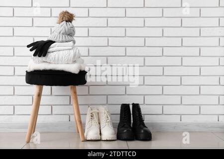 Stack of folded sweaters with hat, gloves on chair and boots near white brick wall Stock Photo