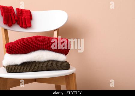 Stack of folded sweaters with gloves on chair against beige background Stock Photo