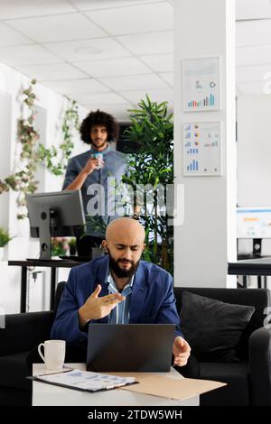 Arab entrepreneur presenting business plan to team during video conference call using laptop. Executive manager discussing product launch in videocall while sitting on couch in coworking space Stock Photo