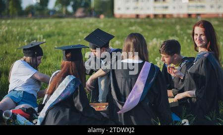 Graduates in black suits eating pizza in a city meadow. Stock Photo