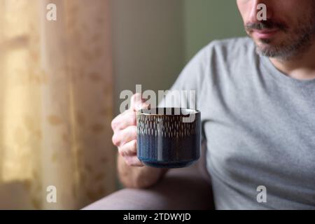 Close up of young caucasian man holding a cup of tea. Bearded man enjoying serene morning with hot tea in his living room at home, relaxing during bre Stock Photo