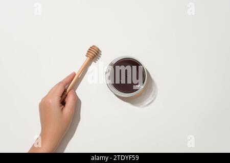 Hand holding a honey drizzle, next to a glass jar of honey on a white background. Honey effectively helps tighten pores. The benefits of honey. Stock Photo