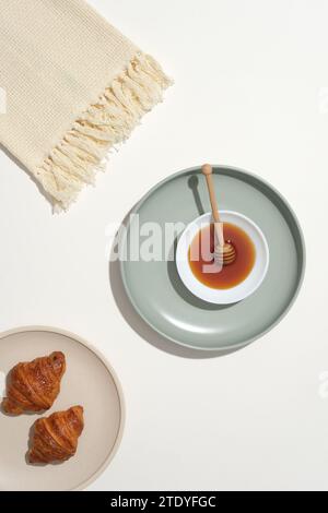 Honey and two croissants are displayed on a round ceramic plate and a beige towel on a white background. Honey contains antioxidants that have antisep Stock Photo