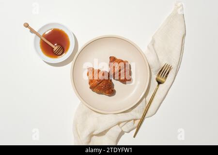 Two croissants are displayed on round ceramic dish, a metal fork, honey and a honey drizzle on a white plate and a cloth towel on a white background. Stock Photo