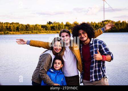 The image depicts a joyful group of four individuals in a picturesque lakeside setting during dusk. The group consists of a young Hispanic woman with long, straight hair, a Caucasian boy in a blue jacket exuding youthful enthusiasm, a Caucasian teenage boy with a bright smile, and a Middle-Eastern young man with curly hair giving a thumbs up. They are all smiling broadly, arms around each other, capturing a moment of carefree happiness. The lake behind them reflects the calming tones of the evening sky, enhancing the feeling of a relaxed and enjoyable outing. Lakeside Bonding: A Group Portrait Stock Photo