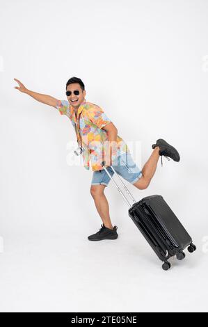 An excited happy Asian man tourist in summer clothes with a luggage is jumping on an isolated white studio background. ready to travel, summer vacatio Stock Photo