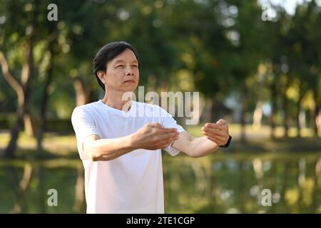 Senior man practicing Chi Kung or Tai Chi gesturing standing against lake during sunrise. Stock Photo