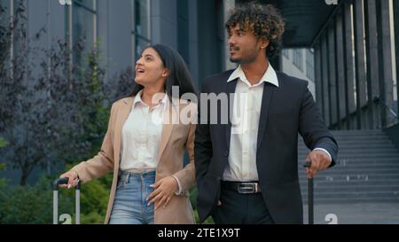 Two diverse multiracial people partners coworkers walking together arriving to city business trip talking pointing direction discuss deal luggage Stock Photo