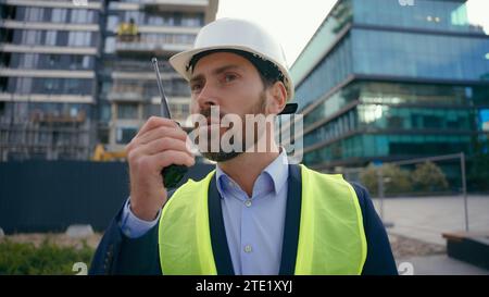 Moving shot Caucasian builder in hard hat helmet and safety vest talking in remote radio outdoors urban building architect engineer contractor Stock Photo