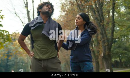 Arabian suffering unwell tired runner man guy feel leg knee pain painful problem injury after running jogging sport Indian girl woman help support Stock Photo