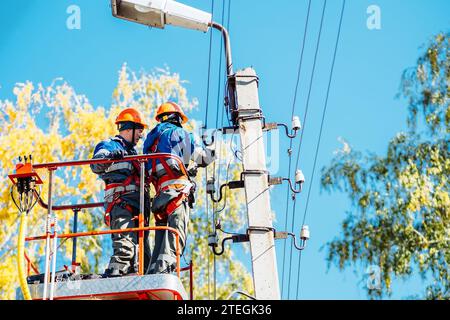 Two professional electricians in hard hats are repairing power lines from cradle of bucket truck. View from below. Electricians change cables on street lighting poles. Stock Photo