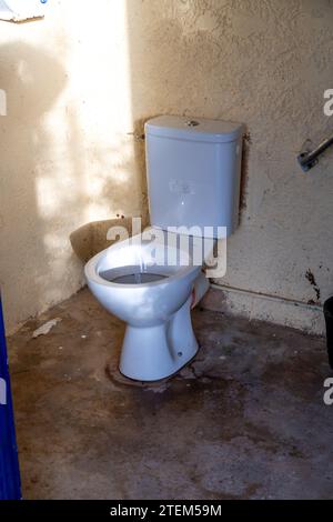 A ceramic toilet with a ceramic cistern in a dirty room without a toilet seat. You can see rays of sunlight and a grab rail in the room. The toilet do Stock Photo