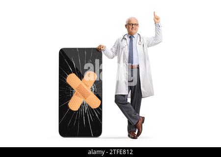 Male doctor leaning on a smartphone with a cracked screen and bandage and pointing up isolated on white background Stock Photo