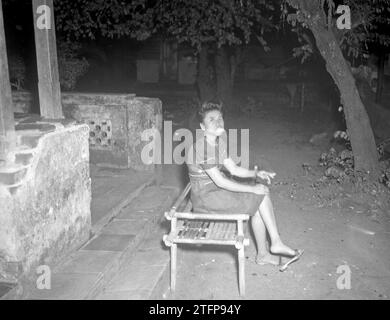 A young woman smokes a cigarette, sitting on a bamboo bench in front of a stone veranda ca. June 22, 1948 Stock Photo