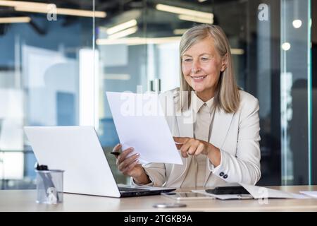 Senior female accountant financier at workplace inside office paperwork, businesswoman satisfied with achievement results checking reports , contracts boss holding paper in hands, working laptop. Stock Photo