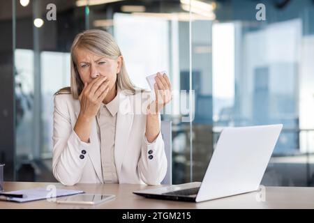 Senior gray-haired woman sick at workplace inside office, businesswoman boss sneezes has cold and allergy, female worker with laptop. Stock Photo