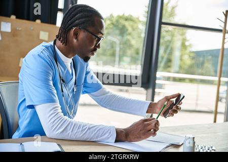 focused african american doctor with glasses taking notes and consulting by phone, telehealth Stock Photo