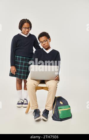 cute african american girl in uniform standing near schoolboy using laptop on grey backdrop Stock Photo
