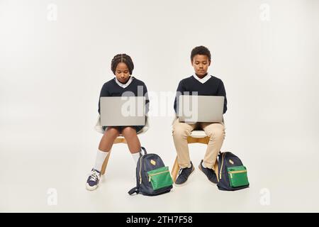 focused african american schoolkids in uniform using laptops and sitting on grey background Stock Photo