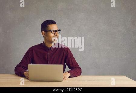 Happy black student sitting at desk with laptop and looking away at copy space background Stock Photo