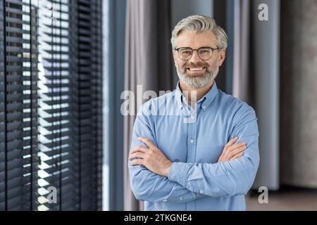Portrait of mature successful businessman boss, senior gray-haired man inside office smiling looking at camera with crossed arms, experienced financier banker accountant in shirt. Stock Photo