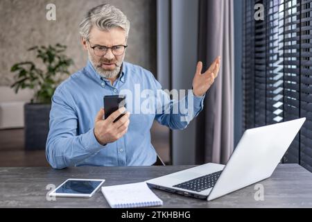 Sad disappointed boss businessman at workplace received online message email on phone, senior mature gray haired man using app on smartphone, inside office with laptop. Stock Photo
