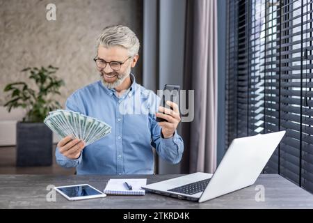 Happy successful financier investor inside office at workplace, senior gray haired businessman holding phone and money cash US dollars in hands, boss celebrating victory and successful achievement. Stock Photo