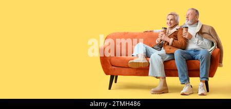Happy mature couple with cups of coffee sitting on sofa against yellow background with space for text Stock Photo