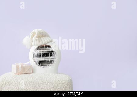 Electric fan heater with hat and gift box on pouf on lilac background. Concept of heating season Stock Photo