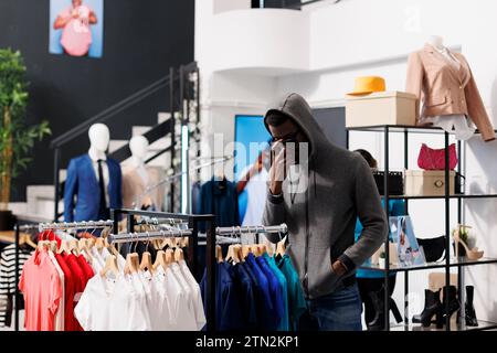 Thief man looking at racks full with casual wear, sneaking stolen trendy clothing in modern boutique. African american robber stealing fashionable merchandise in shopping mall. Burglary concept Stock Photo