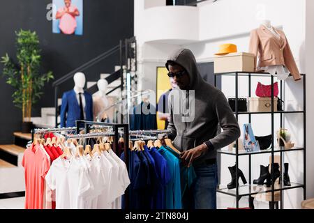 African american thief stealing casual t-shirt, looking around to see if someone watching him in modern boutique. Man robbing clothing store, wearing hood and sunglasses. Crime concept Stock Photo