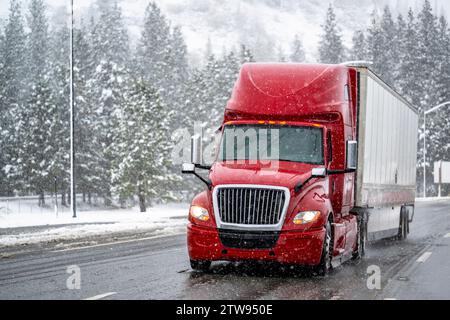 Industrial bright red big rig long hauler semi truck transporting cargo in dry van semi trailer cautiously driving on a dangerous winter highway durin Stock Photo