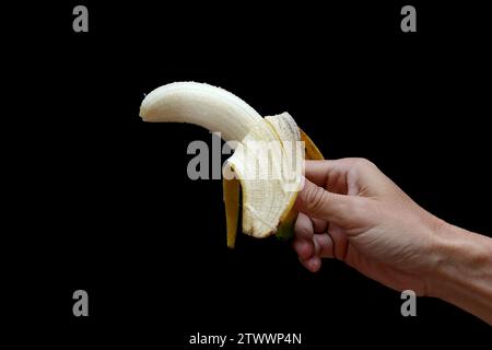 A peeled banana, held up by one hand, against a black background. Stock Photo