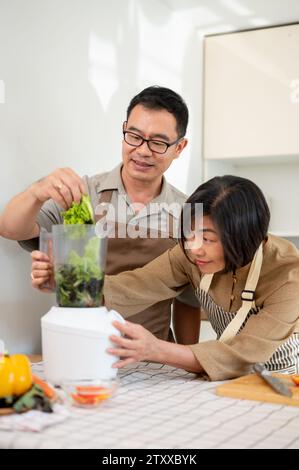 Happy adult Asian couples are enjoying making healthy green smoothie in the kitchen together, using a smoothie blender. Home cooking, family bonding, Stock Photo