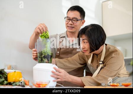 Happy adult Asian couples are enjoying making healthy green smoothie in the kitchen together, using a smoothie blender. Home cooking, family bonding, Stock Photo