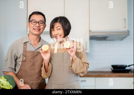 A lovely and happy adult Asian couple is in the kitchen, wearing apron, preparing to cook their dinner together. wife and husband, domestic life, home Stock Photo