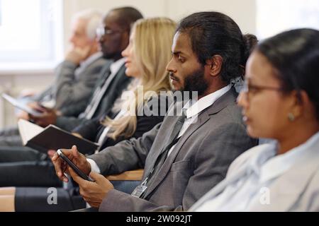 Young businessman in suit using mobile phone while sitting at conference with other people Stock Photo