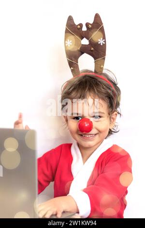 happy boy dressed as Rudolph the reindeer waves his hand while looking at laptop screen, on white background with bokeh. smiling kid. soft focus. vert Stock Photo