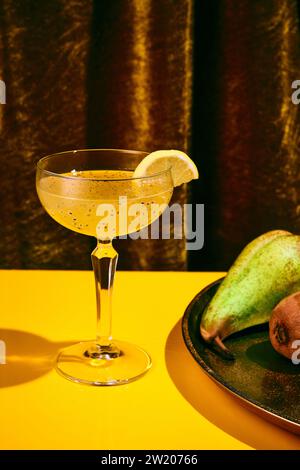 Exotic alcohol drink in tall glass with wide mouth garnished with piece of orange sitting on yellow table with tray of fruit in background. Stock Photo