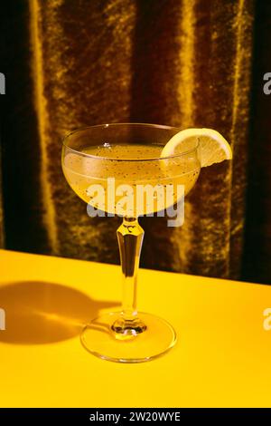 Close up photo of exotic alcohol drink in tall glass with wide mouth garnished with piece of orange sitting on yellow table. Stock Photo