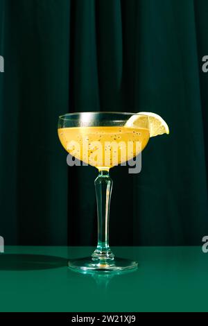 Close up photo of exotic alcohol drink in tall glass with wide mouth garnished with piece of orange sitting on green table. Stock Photo