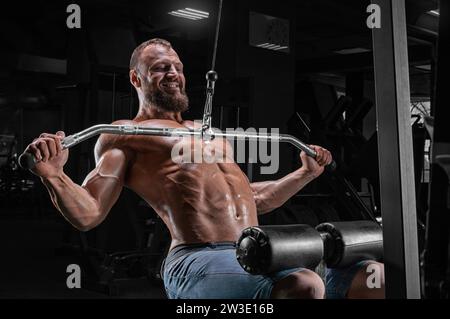 Professional athlete performs an exercise in the gym. Pulls the bar to his chest. Exercise for pumping the back. Mixed media Stock Photo