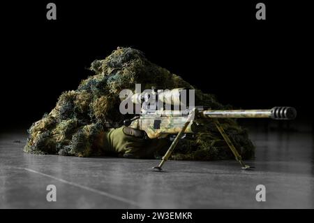 The sniper in disguise lies with a sniper rifle and aims at the telescopic sight. Mixed media Stock Photo