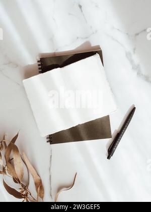 Workspace with black notebook, open blank notebook, pencil and brown leaf on white marble table with shadows. Flat lay, top view office table desk con Stock Photo
